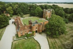 Charming castle wedding venue in Essex and Suffolk, featuring historic architecture, scenic gardens, and elegant surroundings perfect for memorable wedding photography. Aerial view of a historic red brick castle surrounded by lush green trees, manicured lawns, and a gravel driveway, capturing a picturesque setting for weddings in Essex and Suffolk.